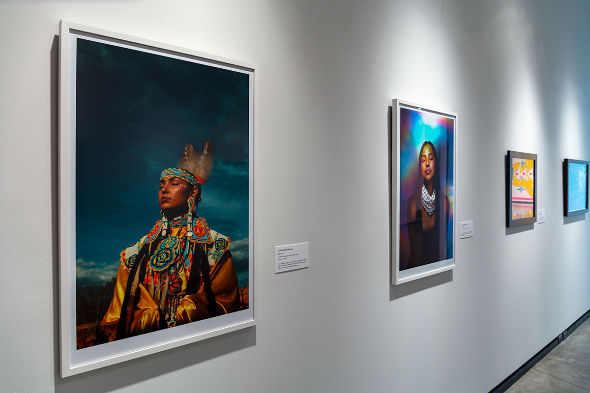 A gallery wall with a row of hanging artworks. The two closest to the viewer are photographs of Native women with their eyes closed, one in traditional dress against a cloudy sky and the other against a background of blue and red color washes