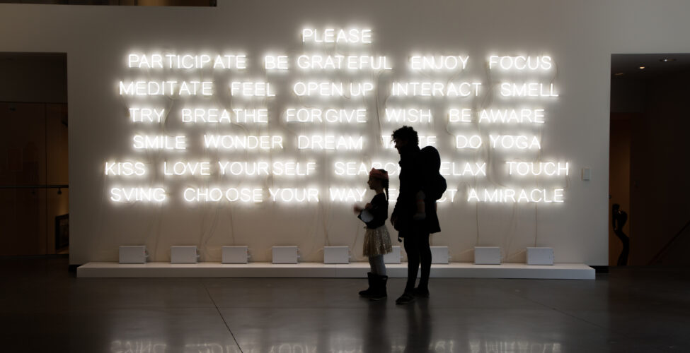 Photo of the silhouette of an adult and a child against a neon word art installation.