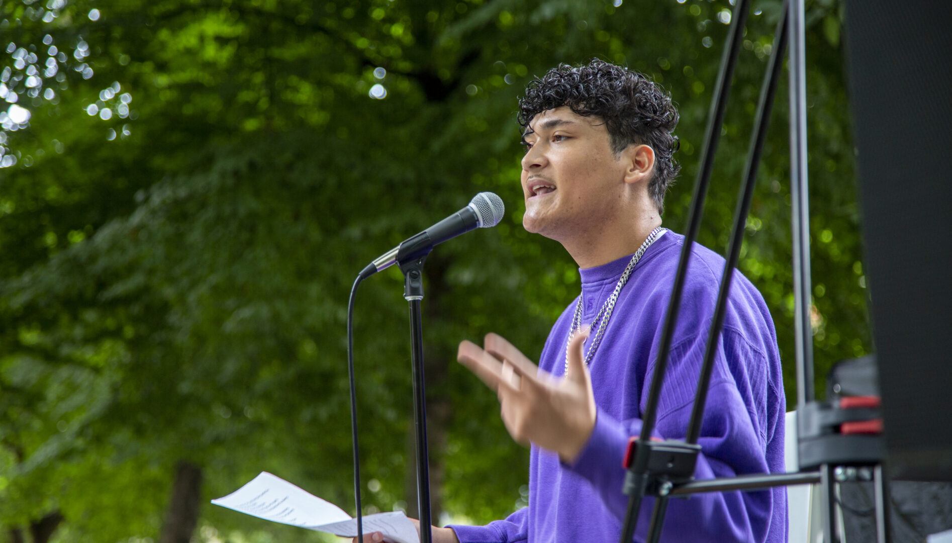 A man in a purple sweatshirt speaking at a microphone under a tree