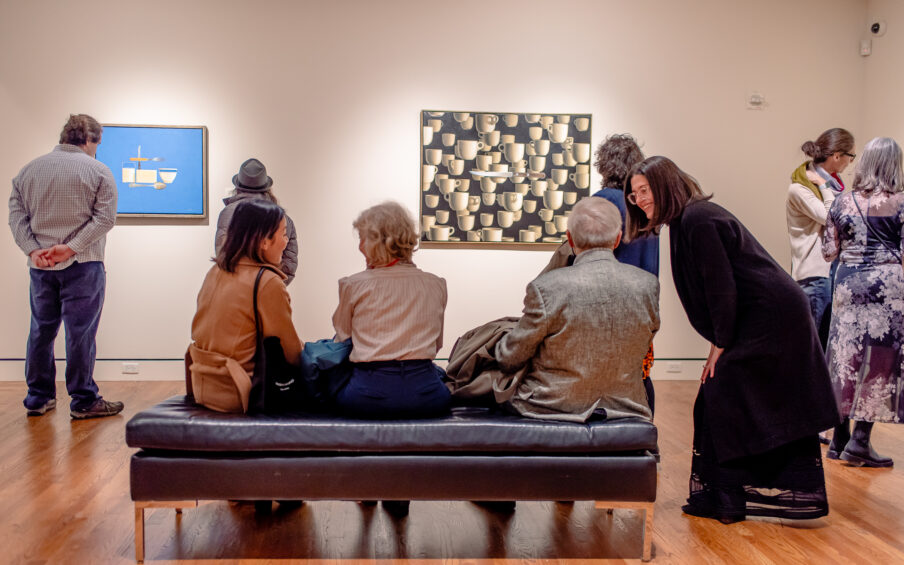 A group of people in a museum gallery, some are sitting on a bench and they are in conversation.