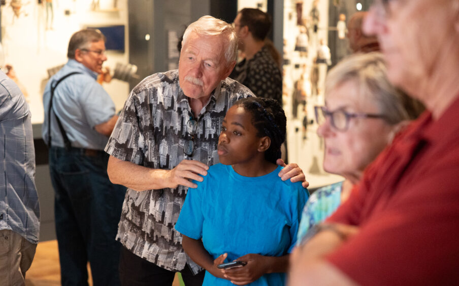 Photo of an older man with his hands on a young boy's shoulders as they both look at art in a gallery.