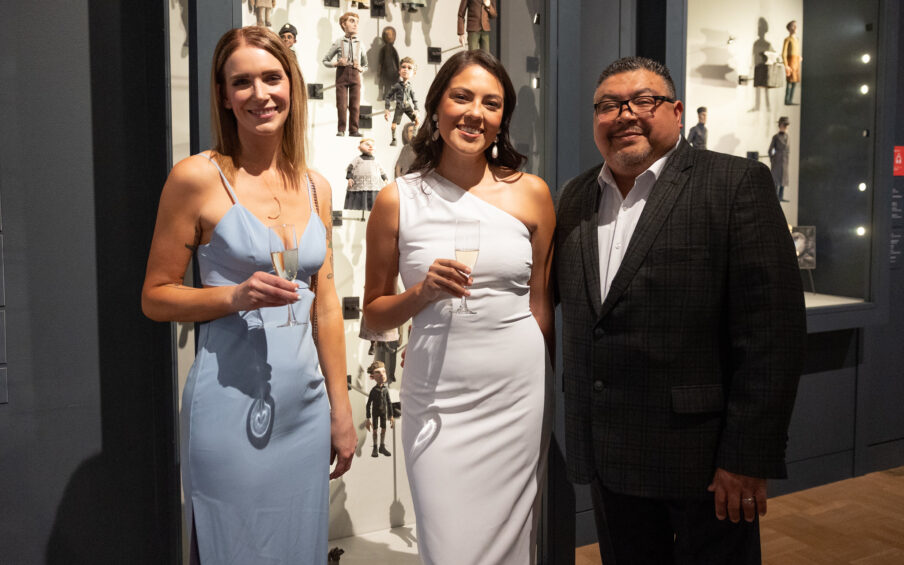 Photo of three people at a semi-formal event in the museum. Two of them are holding champagne glasses.