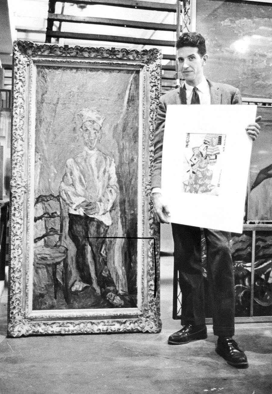 Black and white photo of a man holding a small work on paper standing next to a large framed painting.