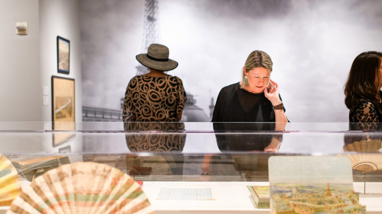 One person examining artwork displayed in a glass case and another person viewing artwork on a wall.