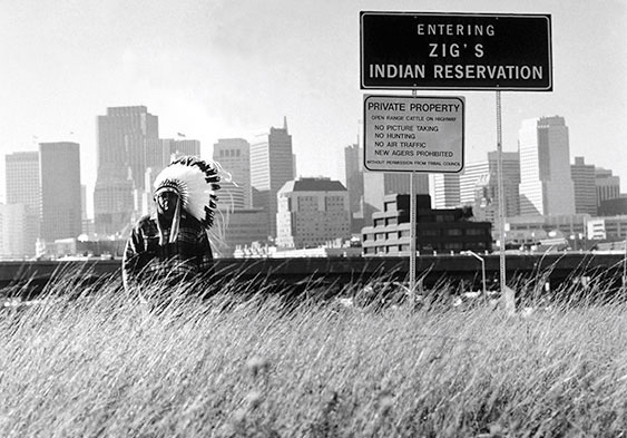 Black and white photo of a Native American man in a feather headdress, standing in a field of grass in front of a cityscape. A sign on the right says "Entering Zig's Indian Reservation"