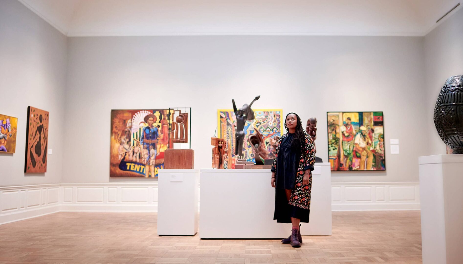 Person standing in gallery space surrounded by paintings and sculptures.