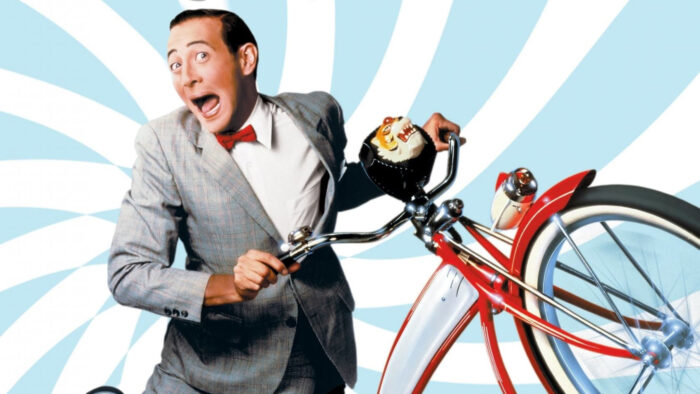 Pee wee Herman in a grey suit, white shirt, and red bow tie, holding on to his red bicycle