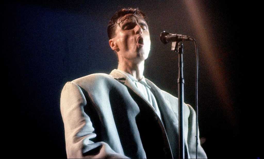 Image of a man at a microphone wearing a very oversized grey suit jacket