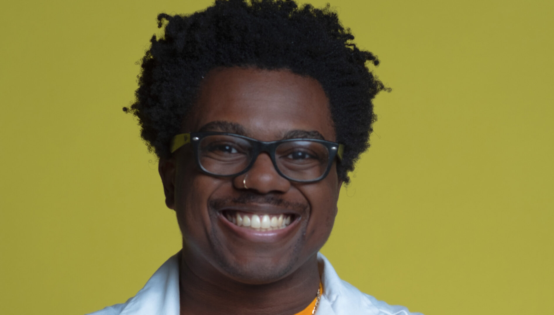 Photograph of a Black person in black eyeglasses and a white lab coat smiling and standing against a green background