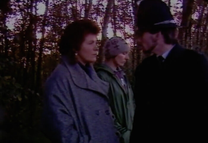 Dark film still of two women and a police officer standing in front of trees