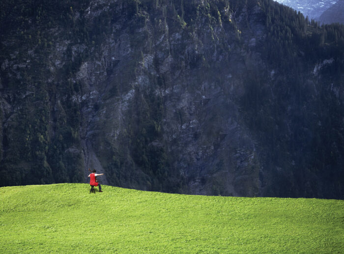 Film still of a person sitting playing a cello on an expanse of green grass at the edge of a cliff facing a mountain