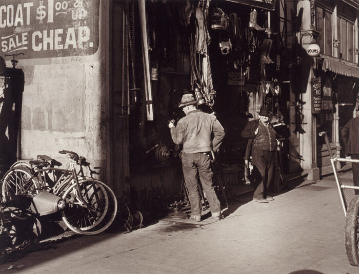 Sepia toned photograph of a a city street scene with one man looking at a wall of tools and one man looking at the camera. Two bicycles are leaning against a wall in the foreground.