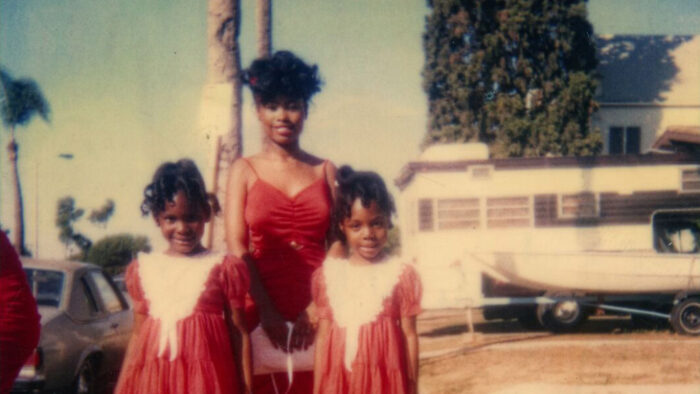 Photograph of a smiling Black woman standing behind two smiling Black girls, all wearing red dresses and standing in front of palm trees outside