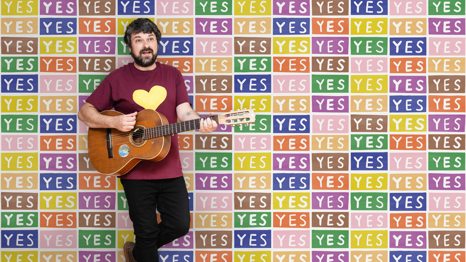 Photograph of a man with a guitar in front of a wall with the word YES repeated
