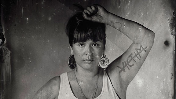 Photo of a Native woman holding one arm up. VICTIM is written and crossed out on her inner bicep.