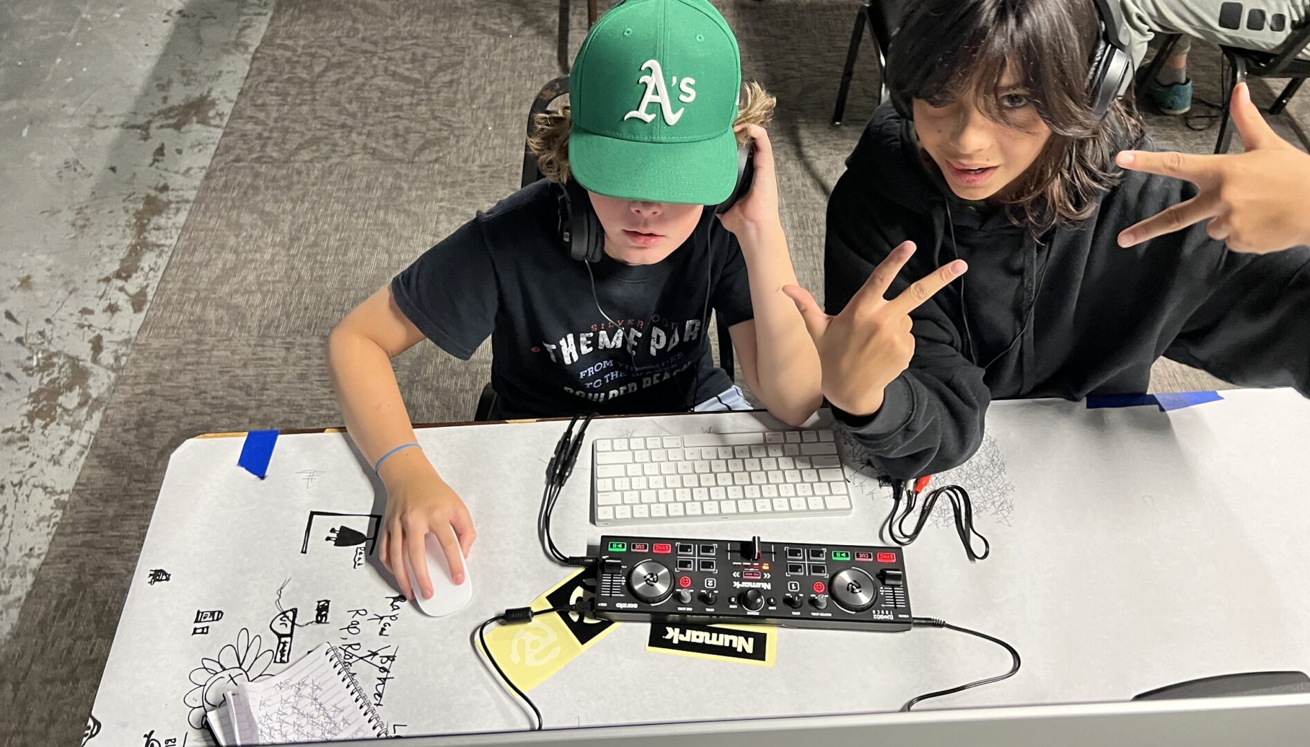 Photo of a kid in a green hat at a mixing board and a kid next to them holding up peace signs