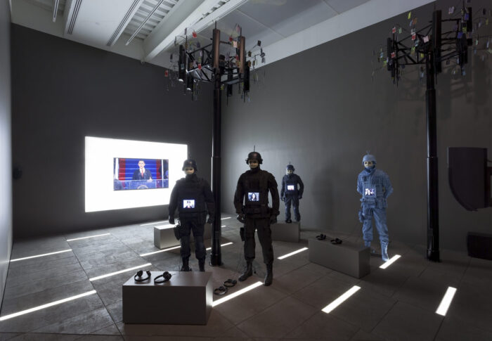 Photo of a dark gallery with four figures in combat gear and a screen showing Barack Obama in the back. There are two towers with what look like surveillance cameras and a headphones are scattered on the floor.