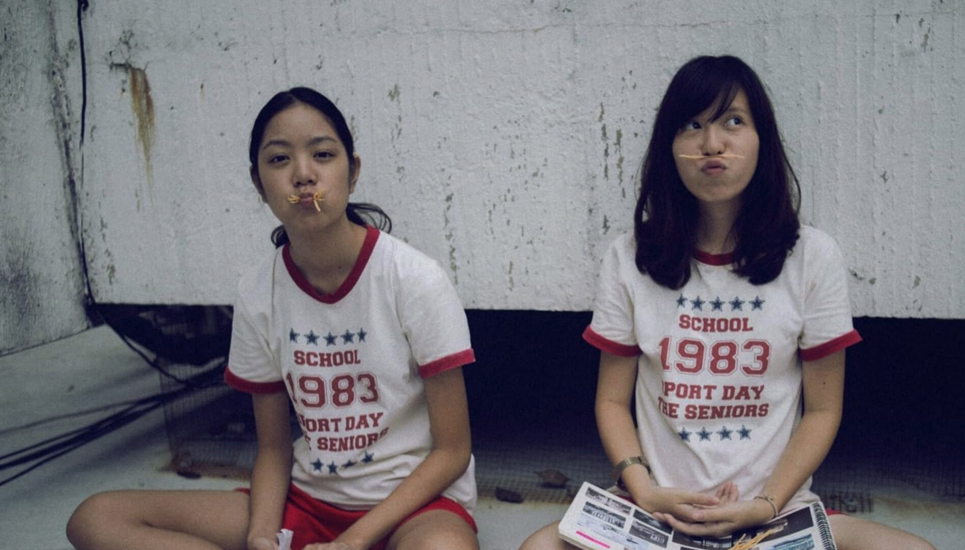 Two young people sitting on a floor, one holding a noodle between their nose and lip, and the other with a mouthful of noodles.