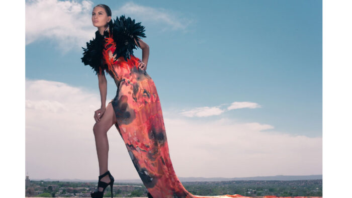 Photograph of a woman wearing a long red and black gown with feather sleeves with a long slit up one side. She is wearing black high heels with one leg out and standing in front of a blue sky and cityscape.
