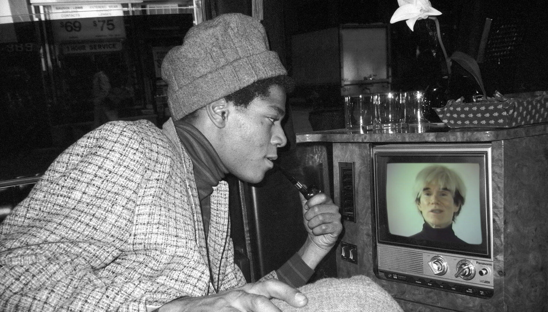 Black and white photograph of Jean-Paul Basquiat looking into a TV screen with a color image of Andy Warhol on it.