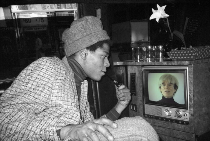 Black and white photograph of Jean-Paul Basquiat looking into a TV screen with a color image of Andy Warhol on it.