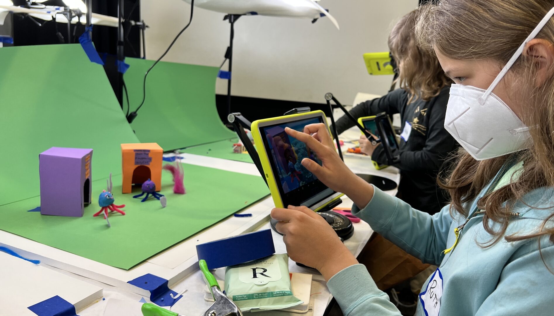 Photo of a child wearing a white mask touching an iPad with a green screen and film set in front of them