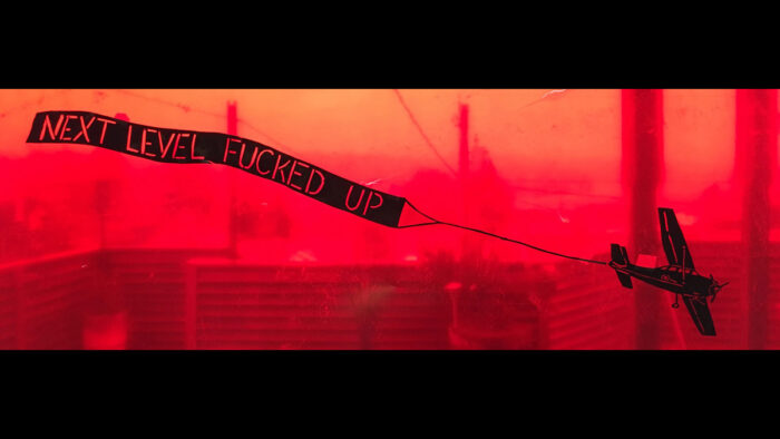 Video still of a red background image with a plane pulling a banner that says "Next level fucked up"