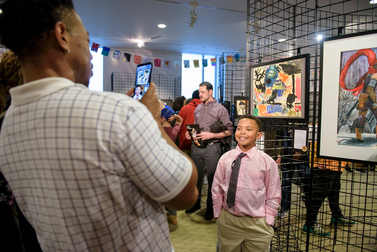 Photograph of a young Black boy in a pink button down shirt and tie standing and smiling in front of his artwork, while a man takes his photo