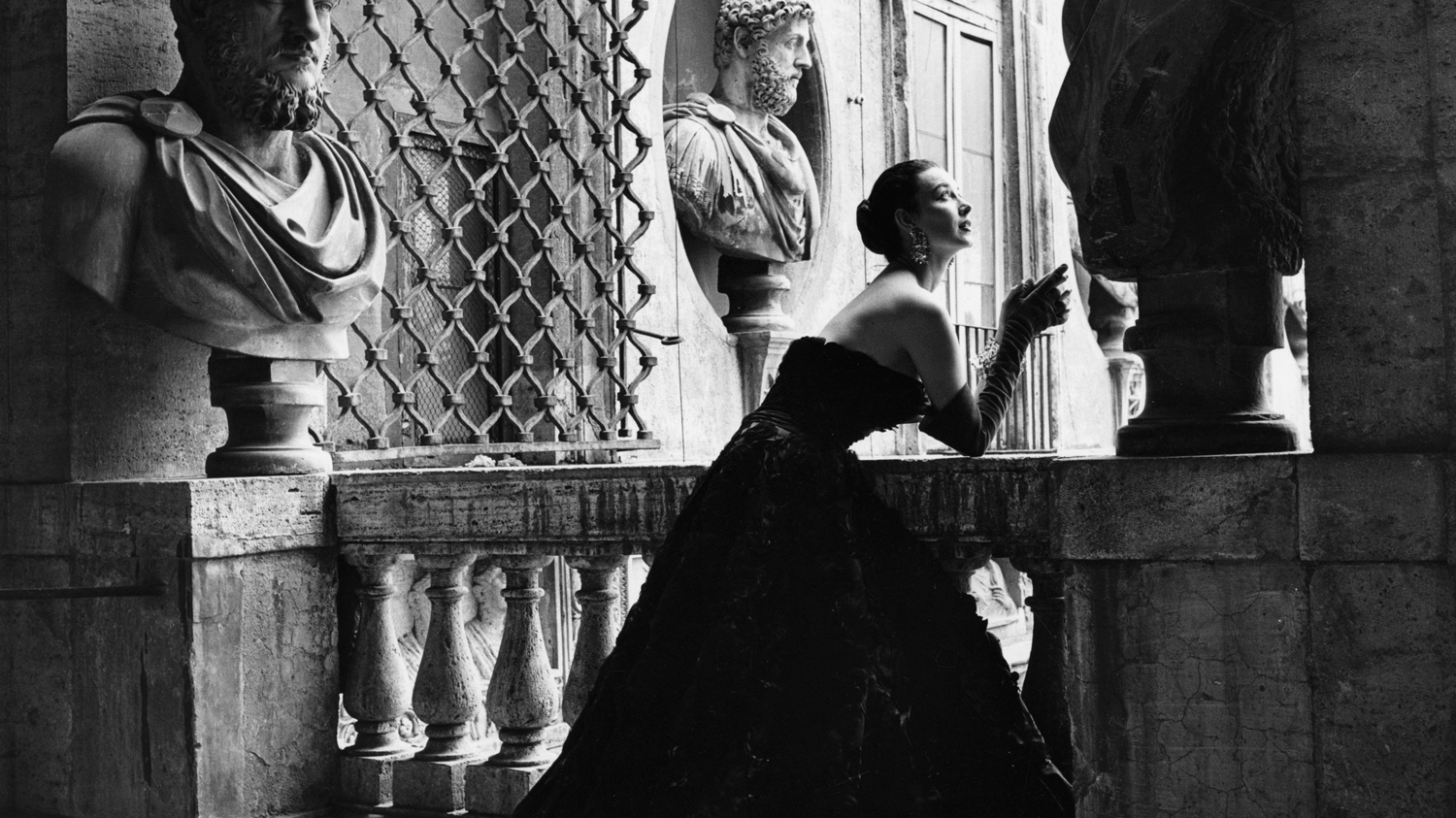 Black and white photo of a woman in a long black sleeveless gown leaning against a stone bannister in a museum-like setting