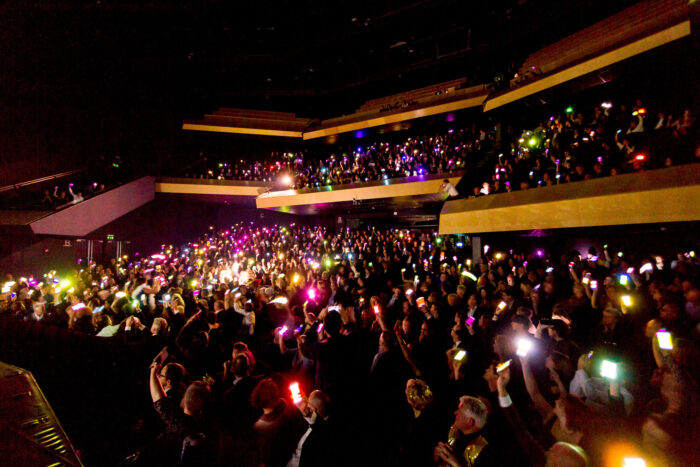 A dark auditorium with hundreds of people holding up their illuminated cellphones
