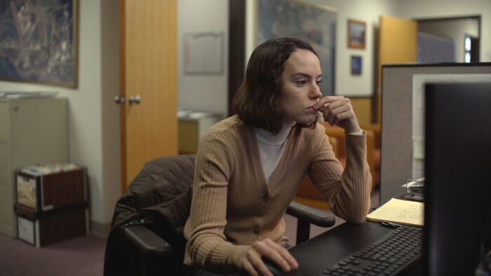 Film still of a woman sitting and looking at a computer in a drab office space