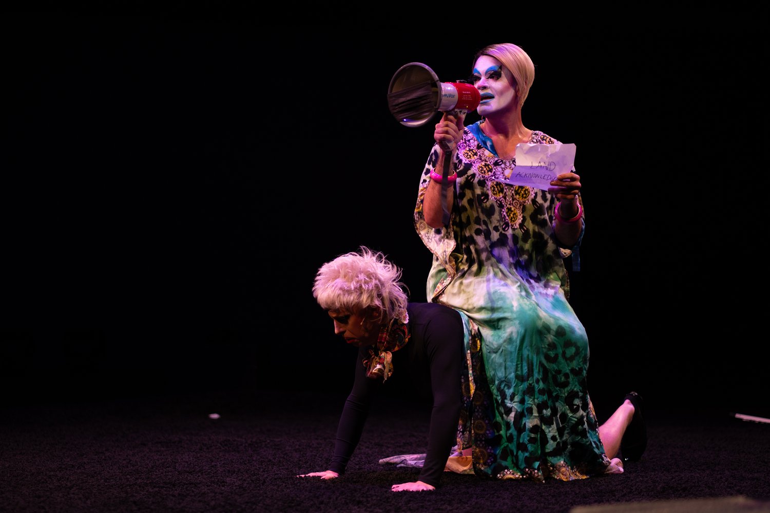 A photo of drag clown Carla Rossi sitting on a dark stage holding a bullhorn. In front of her is another person on their hands and knees facing the audience.