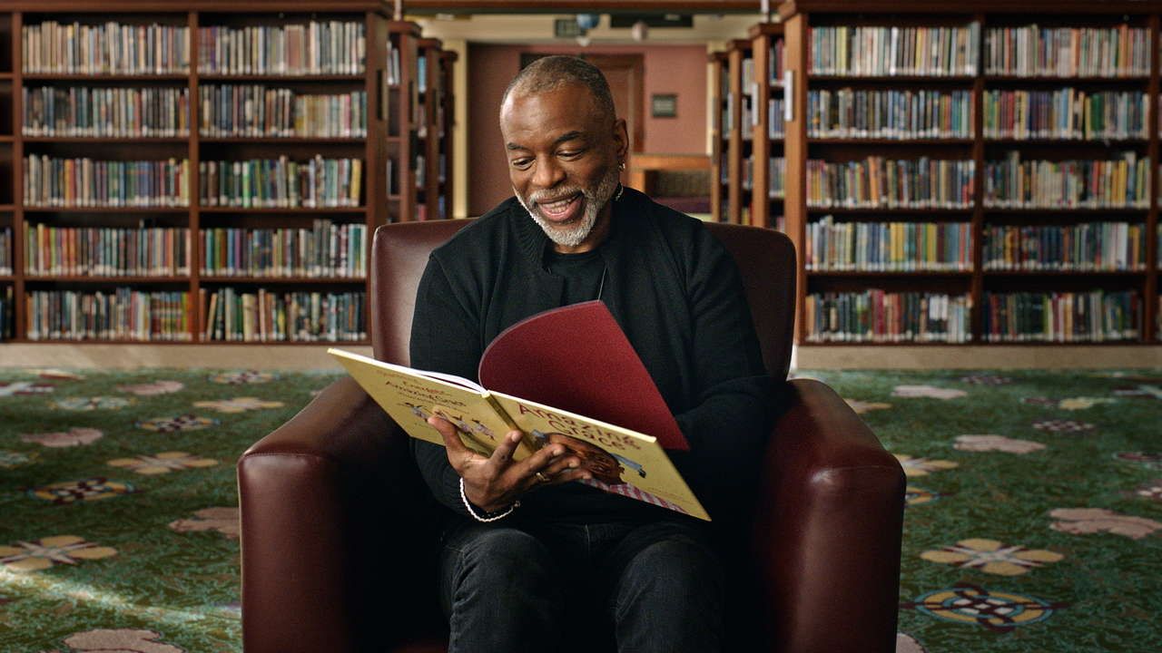 Photo of LeVar Burton sitting in a room with lots of book shelves. He is wearing a black shirt and smiling down at a book.