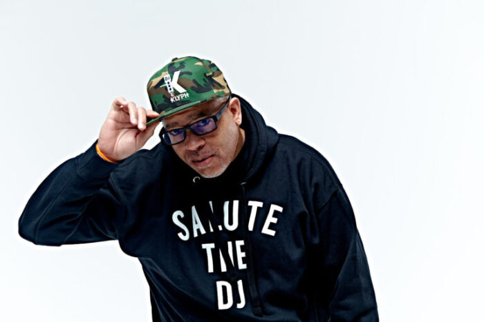Photo of a man with a camouflage baseball hat with a K on it, a black hoodie with "Salute the DJ" written on it, and wearing glasses
