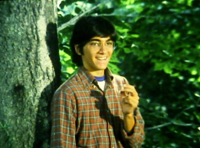 Image of a young man with eyeglasses standing against a tree and smiling, holding a joint