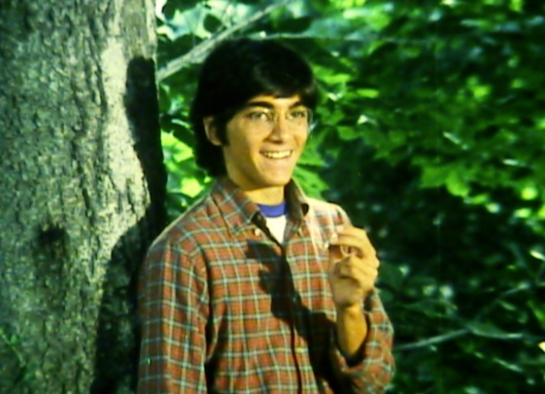 Image of a young man with eyeglasses standing against a tree and smiling, holding a joint