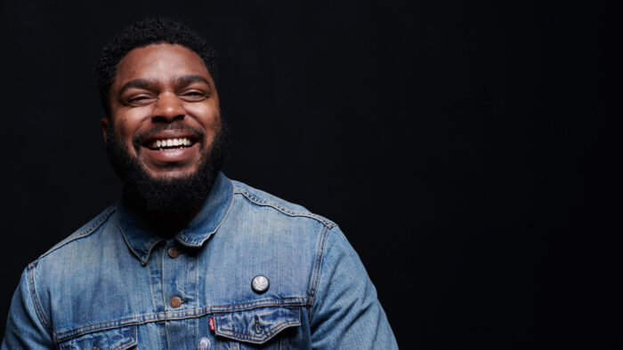 Portrait of a smiling Black man in a denim button down shirt, against a black background