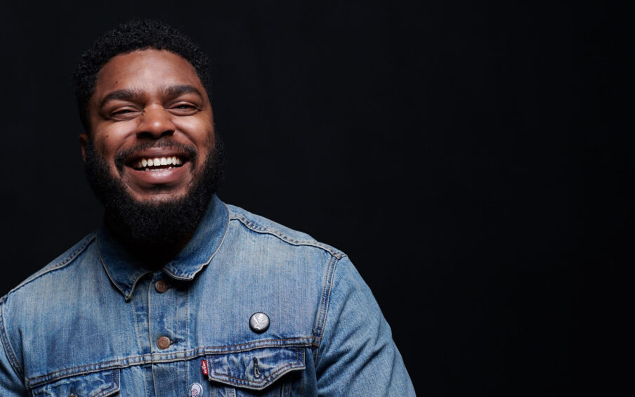 Portrait of a smiling Black man in a denim button down shirt, against a black background