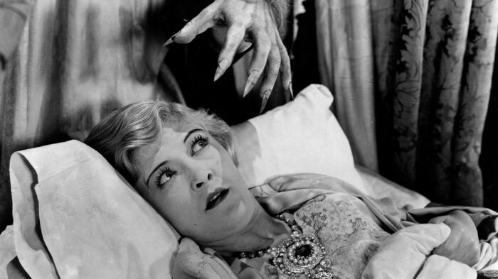 Film still from a black and white movie. A woman is laying on a pillow looking scared with a monster hand reaching down above her