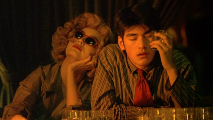 Film still of two people sitting at a dimly lit bar. A woman with blond curly hair, wearing sunglasses, is leaning her head on the shoulder of a man in a tie with his eyes closed, who is holding up a bar glass to his head.