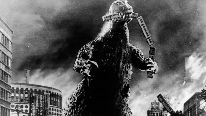 Black and white film still from the original Godzilla movie, of Godzilla standing in a burning city with a train in his mouth