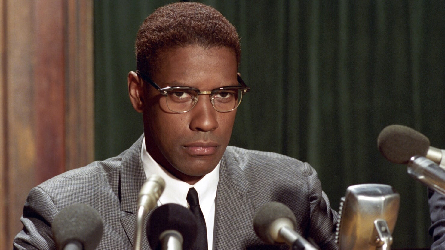 Photo still of Denzel Washington portraying Malcolm X sitting in front of a cluster of microphones