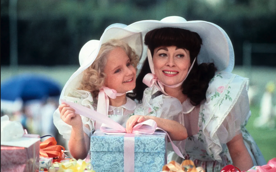 A film still from Mommie Dearest of a mother and her daughter wearing matching wide brimmed white hats in front of a wrapped present and posing for a photo.