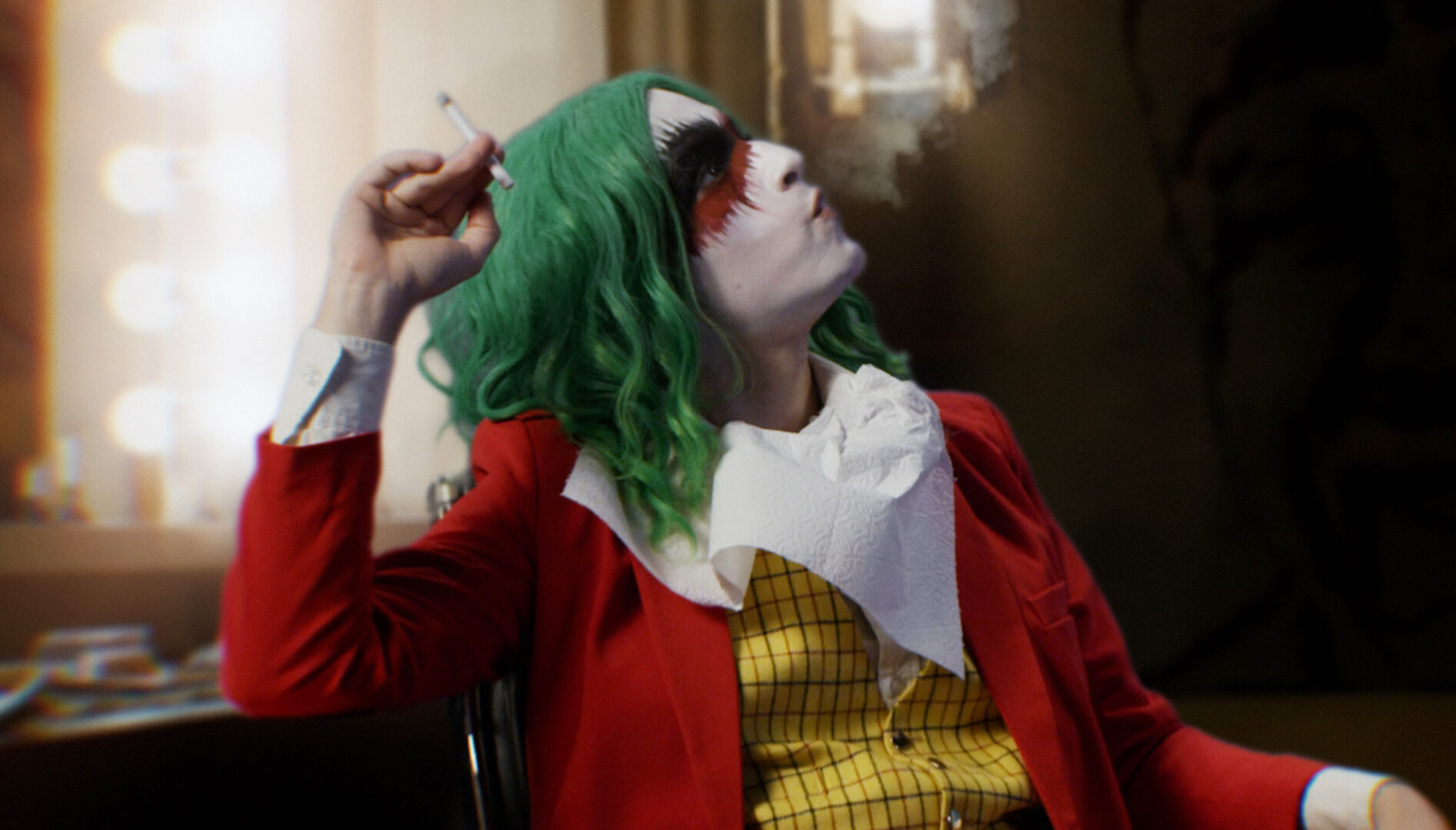 Picture of a person in a Joker's costume sitting in front of a lighted mirror with a cigarette in their hand