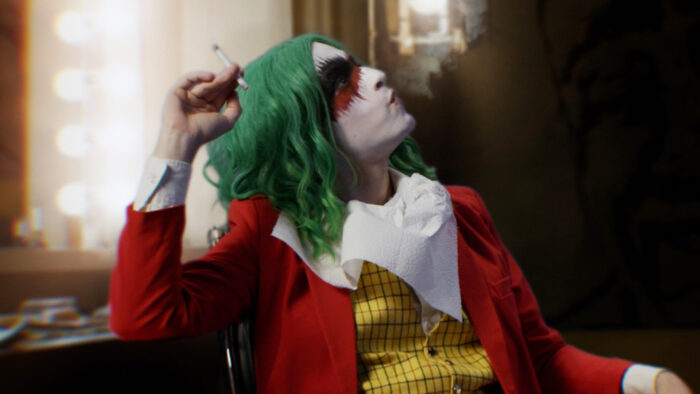 Picture of a person in a Joker's costume sitting in front of a lighted mirror with a cigarette in their hand