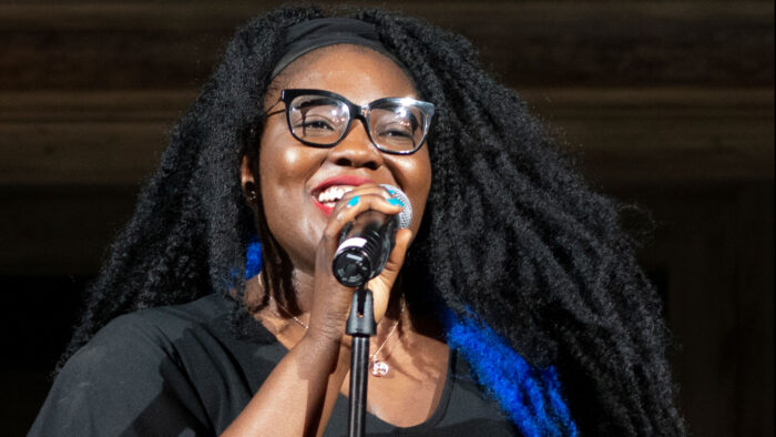 Photo of a smiling Black woman with black eyeglasses and dreadlocks speaking into a microphone