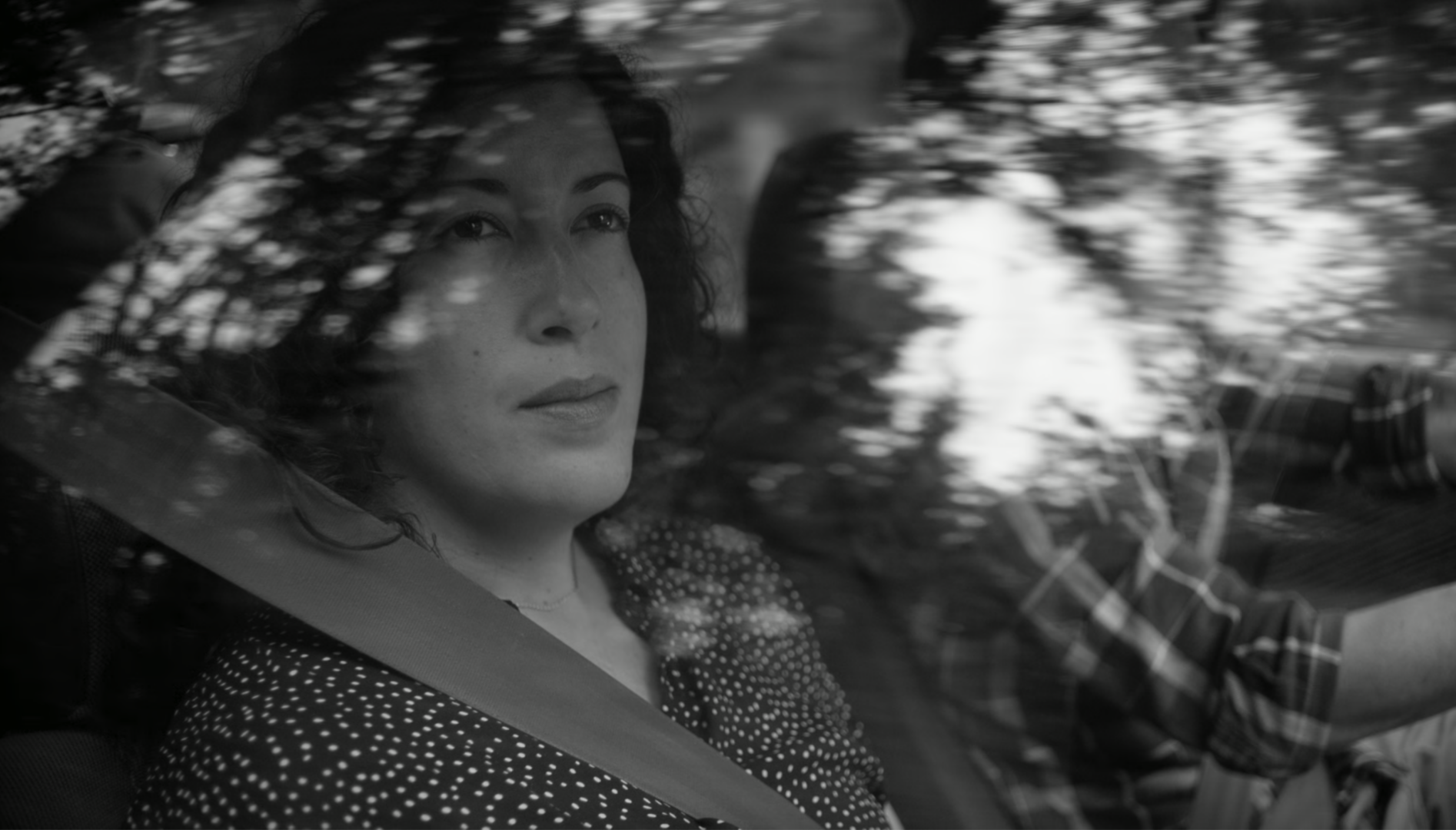 A black and white photo of a woman looking out a car window. The reflection of trees is overlaid on the window.
