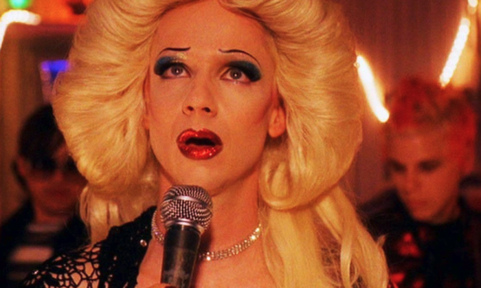 Photo of a person with a blond wig and glamorous makeup singing into a microphone