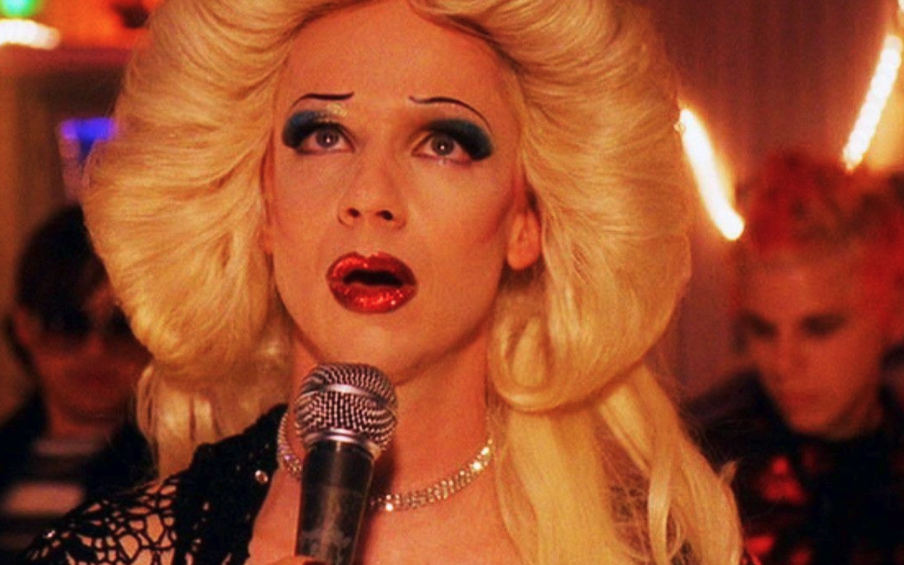 Photo of a person with a blond wig and glamorous makeup singing into a microphone