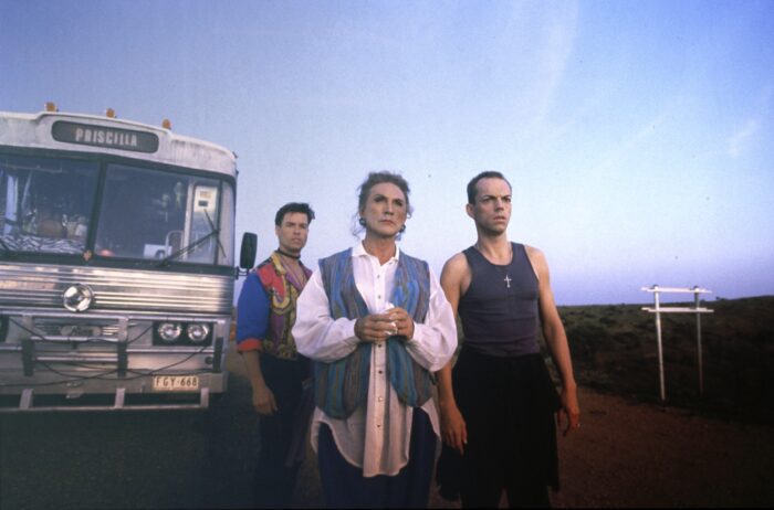 Three men standing in front of a bus that says Priscilla on the top
